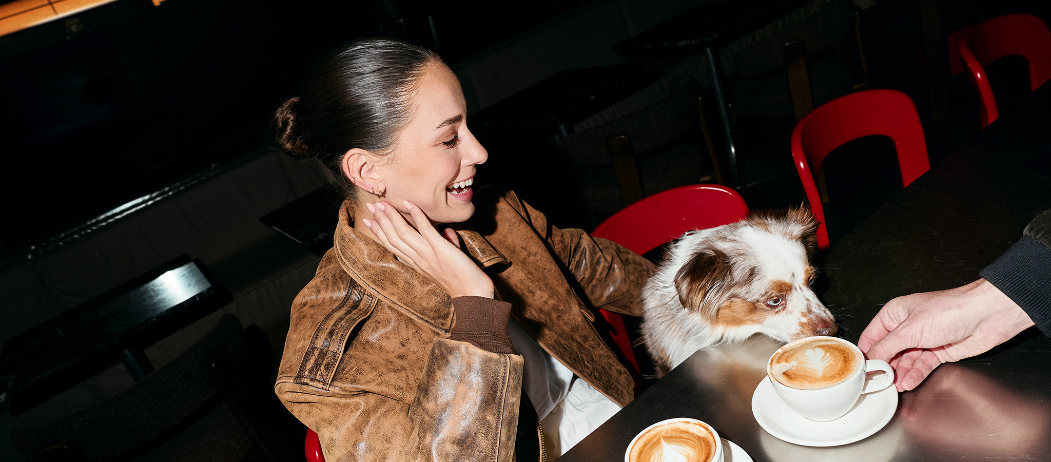 Smiling woman in a leather jacket with her Australian Shepherd at a café table, the dog sniffing a cappuccino