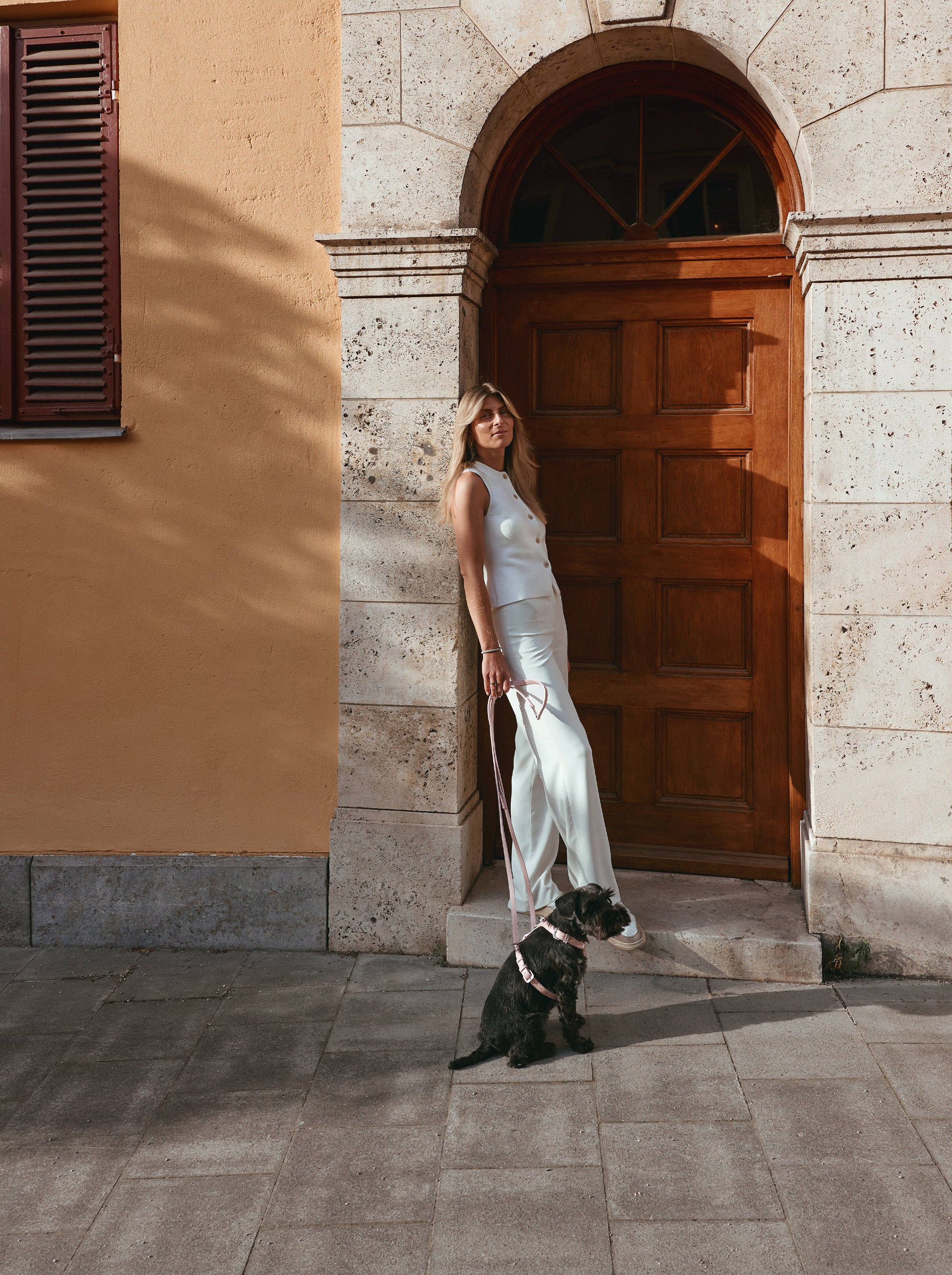 Donna vestita di bianco in una porta illuminata dal sole con il suo Schnauzer al guinzaglio, che indossa la Pettorina Cove e il Guinzaglio Studio Camour color Rosy